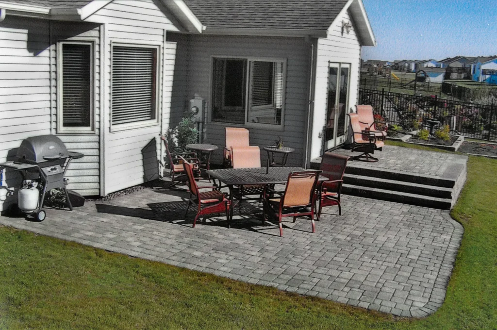 SKM_C451i24121707470 Outdoor patio with stone pavers, dining set, grill, and steps leading to a raised seating area.