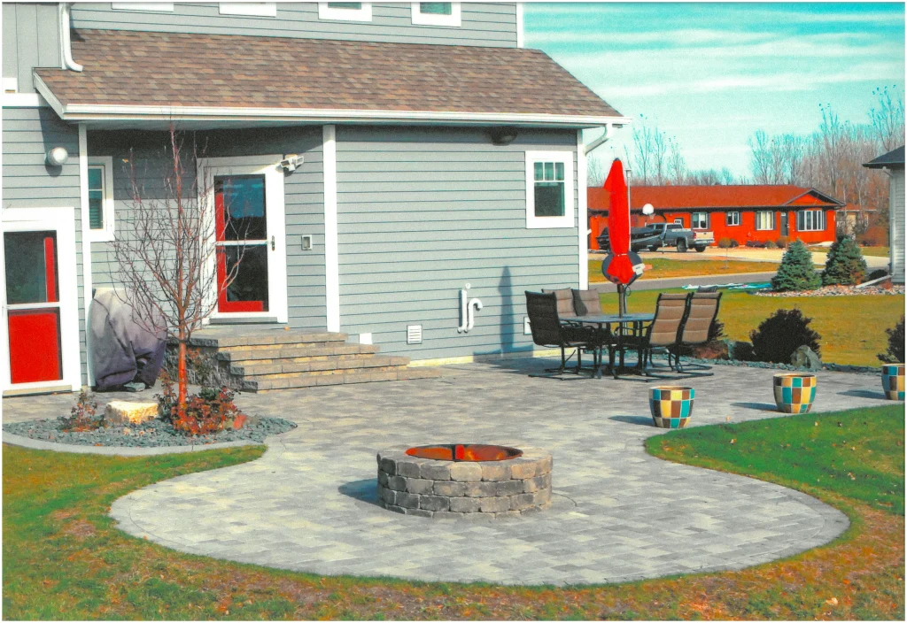 SKM_C451i24121716380_EDIT Stone backyard patio with stone firepit, a tree, shrubs.
