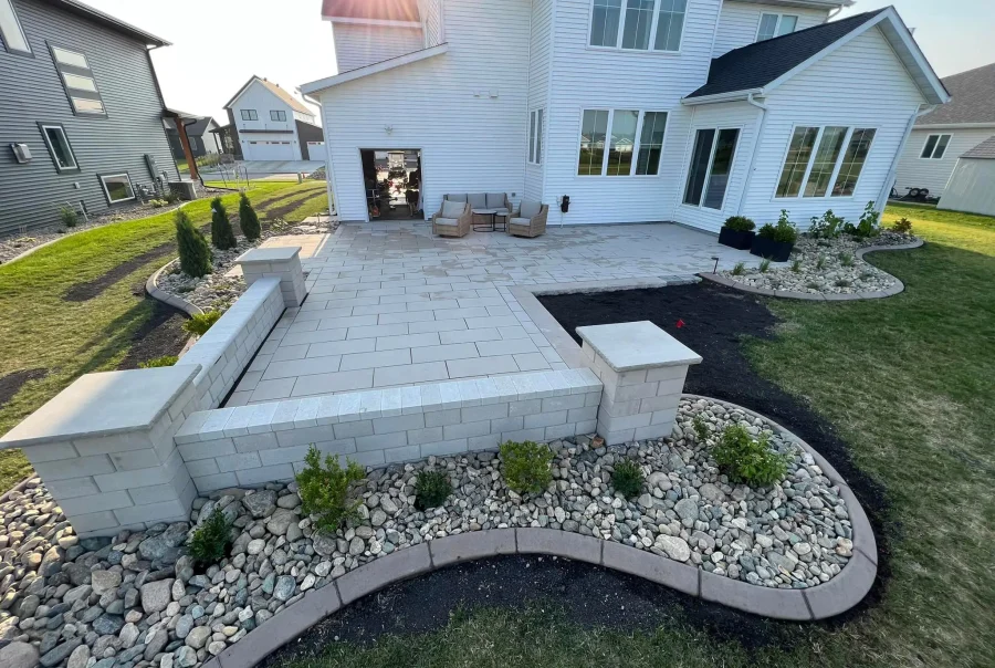 A modern backyard with a tiled patio and landscaped garden, adjacent to a white house.