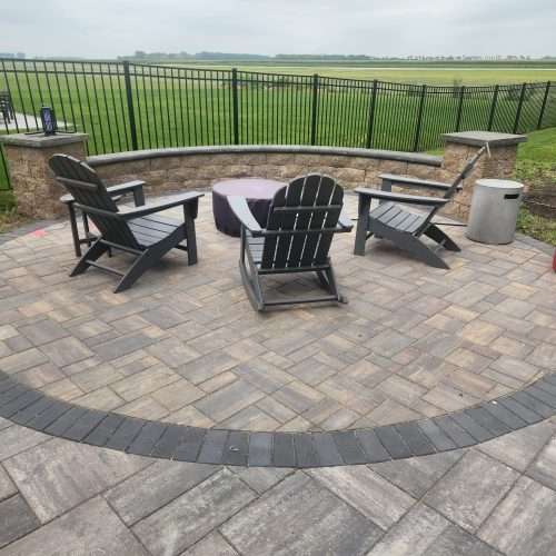 Outdoor patio with Adirondack chairs around a covered fire pit, bordered by a stone wall and metal fence overlooking a green field.