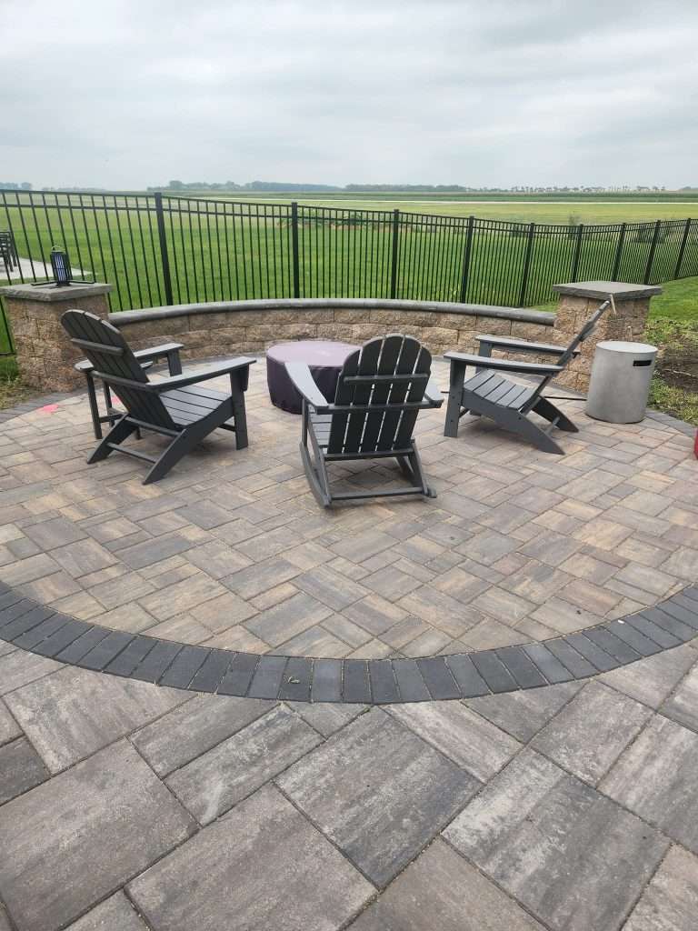 Testimonial_kerriB Outdoor patio with Adirondack chairs around a covered fire pit, bordered by a stone wall and metal fence overlooking a green field.