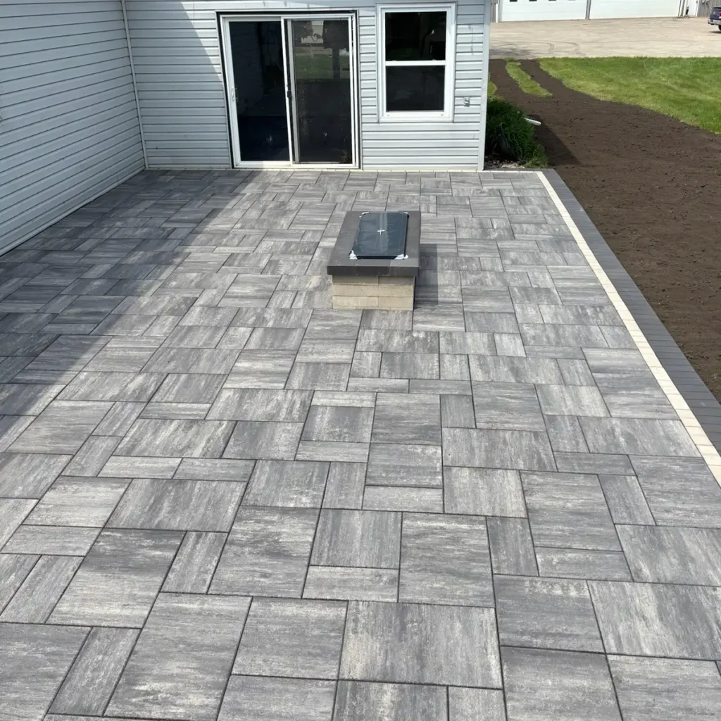 Patio_Fireplace0525-2 A gray stone patio with interlocking paving and a central rectangular fire pit, adjacent to a house.